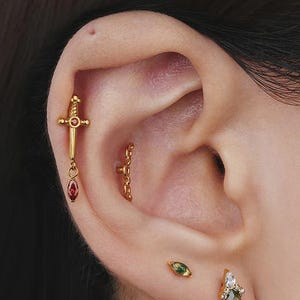 Pode incluir: Uma orelha em close-up adornada com vários brincos dourados. Um brinco em forma de adaga com uma gema vermelha, uma corrente e dois outros brincos pequenos com gemas verdes e brancas. Os brincos estão na pele.