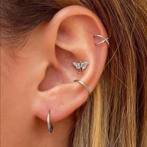May include: Close-up of an ear adorned with multiple silver earrings. A small butterfly stud is centered, with a hoop earring at the lobe and a cross-shaped earring at the upper ear. The ear is set against a blurred background.