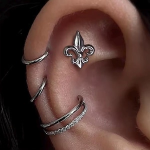May include: Close-up of an ear with multiple piercings. A silver fleur-de-lis stud is centered on the upper ear. Three silver hoops are also visible, one above and two below the stud, with one featuring small, clear stones.