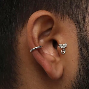 May include: A silver ear cuff and a decorative silver earring are visible on an ear. The ear cuff is a simple, smooth band. The earring features a floral design with a small, red gem at the bottom.