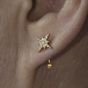 May include: A gold-colored starburst earring with small, clear gemstones set in the points. The earring is attached to a small gold ball that sits behind the ear. The earring is worn on a person's ear.