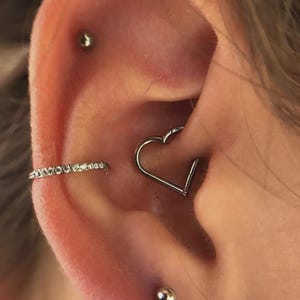 May include: Close-up of an ear with multiple piercings. A silver heart-shaped hoop earring is in the centre, with a small, silver stud above and below. A small, silver hoop earring with clear stones is also visible.