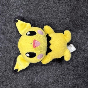 May include: A yellow Pikachu plush toy with black accents on the ears and back. The plush has a pink mouth and cheeks, and black eyes. The toy is lying on a gray surface.