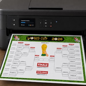 Może przedstawiać: Czarna drukarka drukująca tabelę Mistrzostw Świata 2026 na białej kartce papieru. Tabela zawiera złotą grafikę trofeum i tekst w języku francuskim. Papier leży na drewnianej powierzchni.