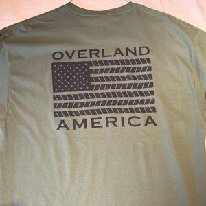 May include: Olive green t-shirt with a graphic of the American flag made of rope and the words "OVERLAND AMERICA" in black. The shirt is made of a soft material and has a crew neck.