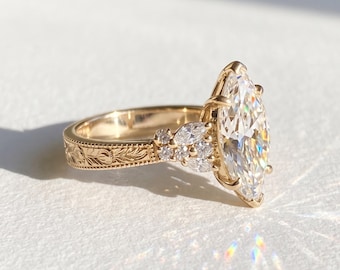 May include: A gold ring featuring a large marquise-cut centre stone held by prongs, flanked by smaller marquise and round-cut accent stones. The band displays an intricate engraved floral pattern along its outer surface, catching the light.