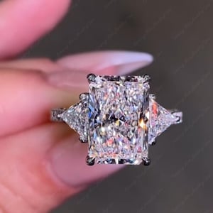 May include: A radiant-cut diamond ring with two triangular side stones. The large, rectangular center stone is the focal point, with the smaller side stones adding sparkle. The ring is set in a silver band, creating a classic and elegant look.