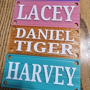 May include: Four colorful nameplates stacked vertically. Each plate is a different color: purple, pink, gold, and teal. White block letters spell out the names: Izzy, Lacey, Daniel Tiger, and Harvey. Each plate has holes.