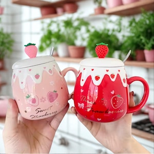May include: Two ceramic mugs with lids, each featuring a strawberry theme. One mug is pink with the text "Sweet Strawberry", and the other is red with the text "Three Strawberries". Both mugs have a strawberry-shaped lid topper and a small spoon.