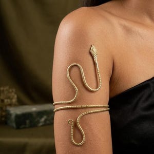 Può includere: Un bracciale da braccio in metallo dorato a forma di serpente avvolto. Il design presenta una superficie testurizzata lungo il corpo del serpente, con una testa dettagliata nella parte superiore e una coda curva nella parte inferiore.