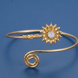 Puede incluir: Una pulsera abierta de tono dorado que presenta un dije en forma de sol en un extremo con un cabujón central de piedra lunar iridiscente. El extremo opuesto de la banda de alambre termina en un diseño de espiral simple y metálico.
