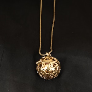 Peut inclure: Un collier avec un pendentif doré. Le pendentif est une sphère ajourée avec un motif complexe. La chaîne est une fine chaîne serpentin dorée. Le collier est présenté sur un fond noir.