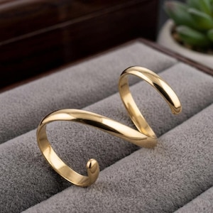 May include: A gold ring with a distinctive, curved design. The ring has a smooth, polished surface and is presented on a grey, velvety surface. The ring's shape is reminiscent of an abstract letter or a stylized wave.