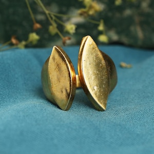 May include: A gold-colored ring with a split design. The ring features two large, leaf-shaped elements that face away from each other. The ring is set against a teal blue background, with hints of greenery in the background.