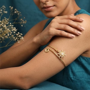 Puede incluir: Brazalete dorado con diseño de sol y perla central. El brazalete tiene un detalle en espiral y se lleva en la parte superior del brazo. La joya se muestra sobre un fondo verde azulado.