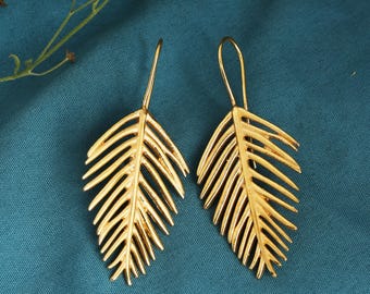 May include: A pair of gold-colored leaf-shaped earrings. The earrings have a detailed vein pattern and a hook closure. The earrings are displayed on a teal-colored background. The earrings are a jewelry item.