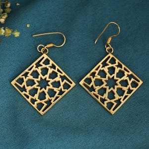 May include: Two gold-coloured earrings with a geometric, openwork design. Each earring is a square shape with a star-like pattern in the centre. The earrings have a hook closure and are displayed on a teal background.