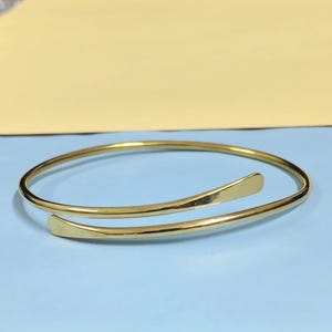 May include: A gold-toned metal bangle bracelet featuring an open, bypass-style design. The bracelet has a smooth, polished finish with two slightly flattened, tapered ends that overlap slightly, creating a continuous, circular, and flexible form.