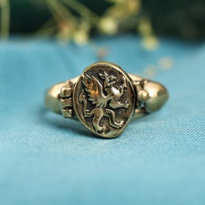 Puede incluir: Un anillo dorado con una cara ovalada que muestra un relieve detallado de una criatura mítica. El anillo tiene una banda texturizada y una estética vintage, sobre un fondo azul.