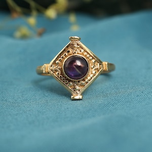 May include: A gold ring with a square, decorative setting. It features a central, round, deep purple gemstone. The ring's design includes detailed patterns around the gemstone and on the band, giving it a vintage or antique appearance.