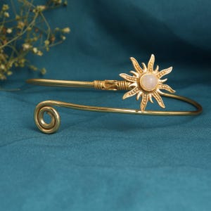 May include: A gold-coloured arm cuff with a sunburst design. The sunburst features a central, round, light-coloured stone. The cuff has a spiral design on one end and a decorative clasp on the other.