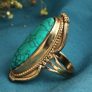 Puede incluir: Anillo dorado con una gran piedra turquesa ovalada con vetas negras. El anillo tiene una banda y un engaste detallados y texturizados, con un acabado pulido. La piedra turquesa es de un color verde azulado vibrante.