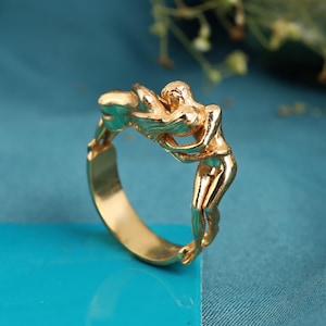 May include: A gold-colored ring featuring two figures embracing. The ring's design depicts the figures in a continuous loop, with intricate details. The ring is set against a blue background, highlighting its metallic sheen.