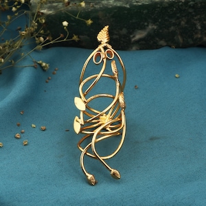 May include: A gold-colored arm cuff with a leaf and vine design. The cuff features multiple loops and leaf accents, creating an elegant and nature-inspired aesthetic. The cuff is displayed on a teal-colored surface.