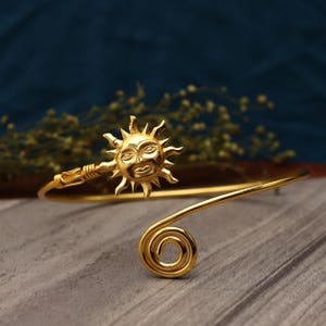 May include: A gold-toned open cuff bracelet featuring a central sun motif with a detailed face and radiating rays on one end, and a simple spiral coil design on the opposite end. The metal band is smooth and polished with a bright yellow finish.