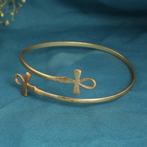 May include: A gold-colored arm cuff with an open design. The cuff features two ankh symbols, one at each end. The ankhs are detailed with a cross at the top and a loop below. The cuff is displayed on a teal blue surface.