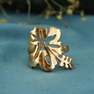 May include: A gold-colored ring in the shape of a stylized flower. The ring features intricate cut-out details and a delicate, leaf-like accent. The ring is set against a blue background.