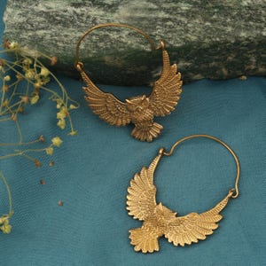Könnte beinhalten: Ein Paar goldfarbene Creolen mit einem Eulen-Design. Die Ohrringe zeigen ausgebreitete Flügel und einen kreisförmigen Bügel. Die Federn der Eule sind detailliert. Die Ohrringe liegen auf einem blauen Untergrund.