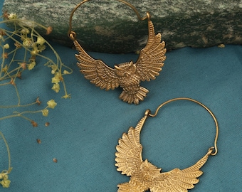 May include: A pair of gold-colored hoop earrings featuring an owl design with outstretched wings. The earrings are detailed with feathers and have a circular hoop for the ear. The earrings are displayed on a blue surface.