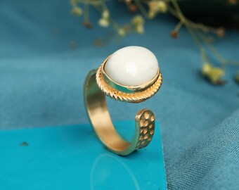 May include: A gold-colored ring with an oval, white gemstone. The ring has a textured band and a decorative setting around the stone. The ring is open at the bottom, with a hammered texture on the band.