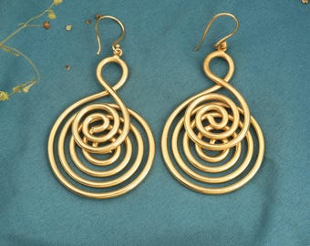 May include: A pair of gold-colored spiral earrings. Each earring features a large, open spiral design with multiple concentric circles, creating a layered effect. The earrings have a hook closure. The background is a teal color.