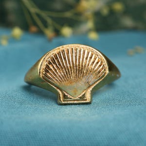 Puede incluir: Un anillo dorado con un diseño de concha. El anillo presenta un motivo de concha detallado en forma de abanico sobre una base rectangular. El anillo está sobre un fondo azul.