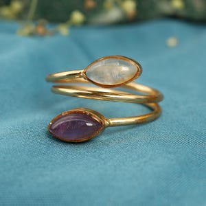 Puede incluir: Un anillo en espiral dorado con dos piedras preciosas en forma de marquesa. Una gema es azul pálido y la otra morada oscura. El anillo está sobre un fondo de tela color turquesa.