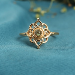 May include: A gold ring with an openwork floral design. The ring has a central, round, pale green gemstone. The band is thin, and the ring is set against a teal blue background.