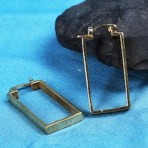 May include: A pair of gold-toned hoop earrings featuring a rectangular frame design. The earrings have a hinged closure mechanism at the top and a smooth, polished metallic finish, showcasing a clean geometric shape and a simple, open structure.