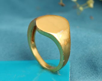 May include: A gold-colored signet ring with a flat, oval face. The ring has a smooth, polished finish and a classic, minimalist design. The ring is set against a teal background.