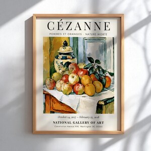 May include: A framed exhibition poster featuring Paul Cézanne's Still Life with Apples and Oranges. The print displays a decorative jar and assorted fruit on a white cloth, with text reading CÉZANNE, POMMES ET ORANGES, and NATIONAL GALLERY OF ART.