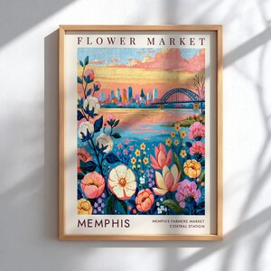 Peut inclure: Une impression d'art encadrée intitulée FLOWER MARKET MEMPHIS représentant une illustration colorée d'un paysage urbain au bord d'une rivière avec un pont, un ciel au coucher du soleil et des fleurs roses, jaunes et blanches au premier plan.