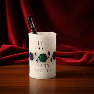 Peut inclure: Un porte-stylo cylindrique en pierre blanche avec des motifs découpés complexes et une bande horizontale de fleurs incrustées dans des tons de bleu, vert et rouge. Un stylo noir est inséré dans l'ouverture supérieure du récipient.