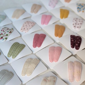 May include: A collection of hair clips in various colors and patterns, including pink, yellow, and floral designs. Each clip is displayed on a white card, creating a visually appealing arrangement. The clips are made of fabric and are likely for styling hair.
