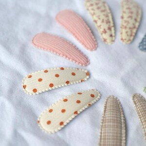 May include: A collection of fabric hair clips in various patterns and colors. The clips include pink ribbed, cream with orange polka dots, floral, and checkered designs. They are arranged on a white, textured surface.