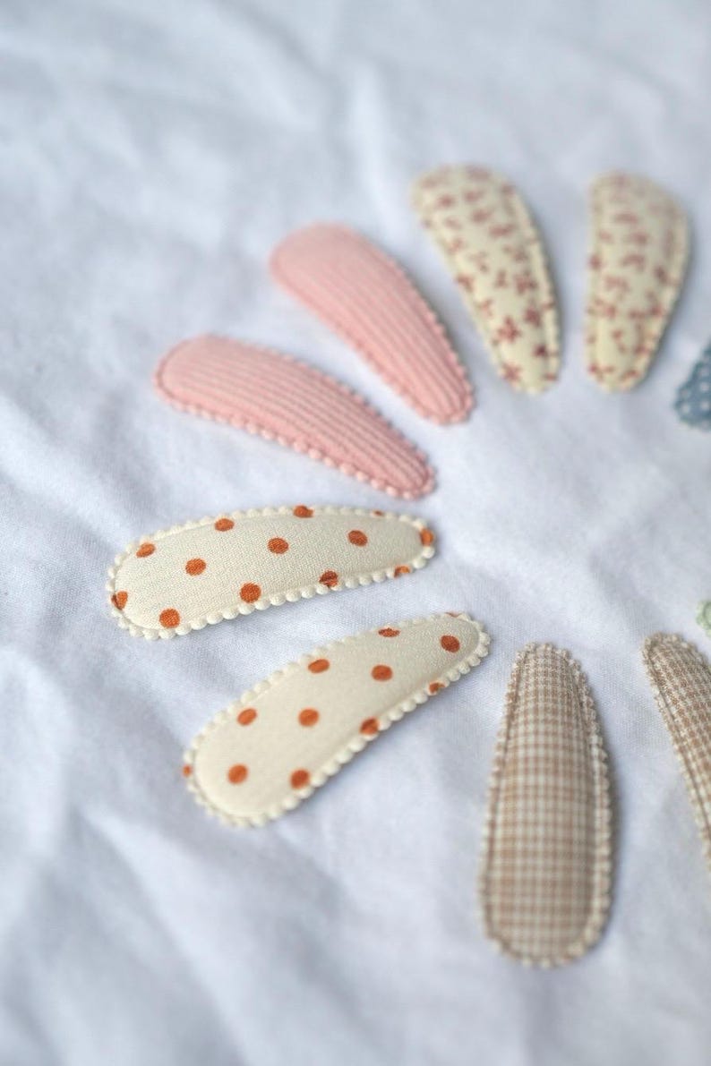 May include: A collection of fabric hair clips in various patterns and colors. The clips include pink ribbed, cream with orange polka dots, floral, and checkered designs. They are arranged on a white, textured surface.