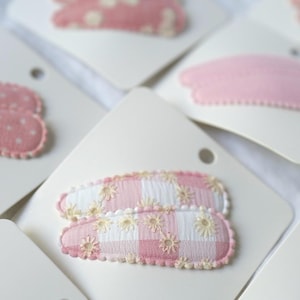 May include: Close-up of several hair clips on white cards. The clips feature various pink patterns, including polka dots, solid pink, and gingham with embroidered flowers. The edges of the clips are scalloped.
