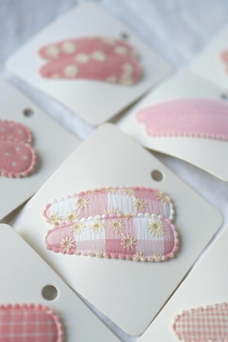 May include: Close-up of several hair clips on white cards. The clips feature various pink patterns, including polka dots, solid pink, and gingham with embroidered flowers. The edges of the clips are scalloped.