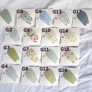 May include: A collection of sixteen fabric hair clips, each attached to a white card. The clips feature various patterns, including floral, gingham, and polka dots, in shades of blue, green, and cream.
