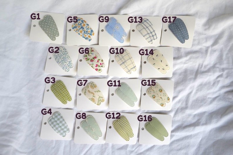May include: A collection of sixteen fabric hair clips, each attached to a white card. The clips feature various patterns, including floral, gingham, and polka dots, in shades of blue, green, and cream.